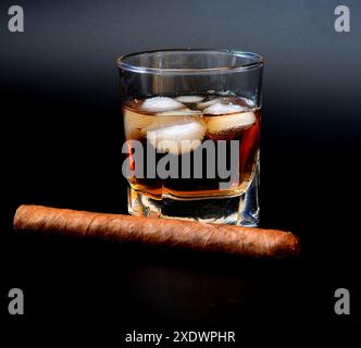 A large Cuban cigar and a glass of burning whiskey with ice on a black background. Close-up. Stock Photo