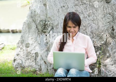 Asian woman sitting green park using laptop computer. Woman working on laptop happy entrepreneur business using notebook with hands typing on keyboard Stock Photo
