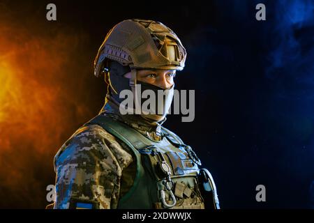 Portrait of a Ukrainian armed forces soldier in full equipment against yellow blue smoke on a dark background, concept of military conflict in Ukraine Stock Photo