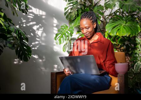 Calm black woman among home garden. Happy african american girl with portable laptop in greenhouse Stock Photo