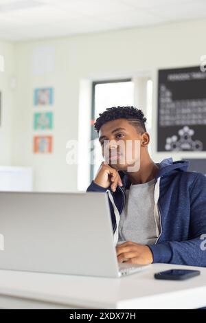 In school, male student using laptop and thinking in classroom, smartphone on desk, copy space Stock Photo