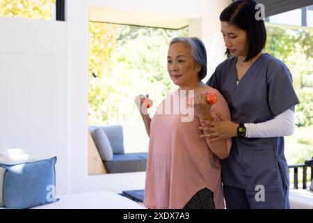 Female physical therapist assisting senior woman with dumbbell exercises at home Stock Photo