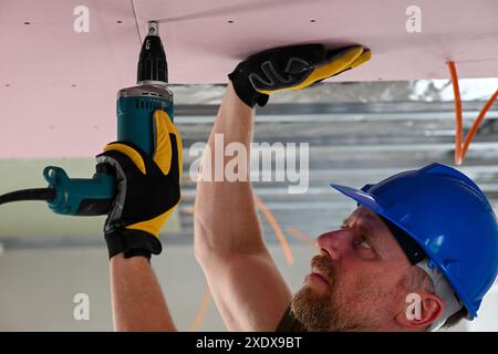 Construction worker wearing protective gear using power drill to install drywall on ceiling in new home construction. Stock Photo