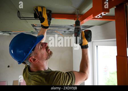 Construction worker wearing a hardhat and safety glasses using a cordless drill to install drywall to the ceiling. Stock Photo