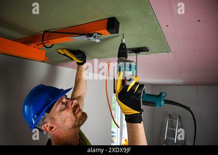 Construction worker wearing protective gear using power drill to install drywall on ceiling in new home construction. Stock Photo