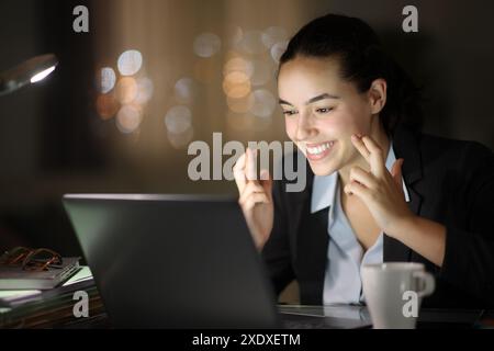 Happy executive working in the night crossing fingers checking laptop Stock Photo