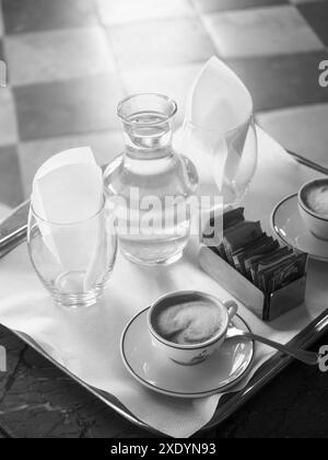 Venice, Italy - June 30th 20220 Black and white photo of a tray holding a steaming cup of coffee, a water carafe, sugar packets and empty glasses in a Stock Photo