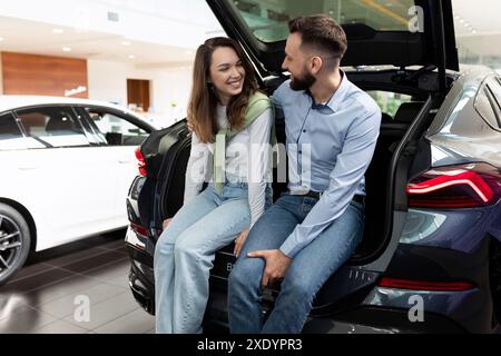 Happily young couple sitting in car trunk dangling legs in car dealership Stock Photo
