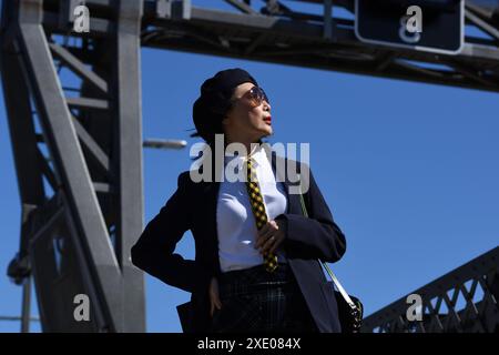 Woman in a white shirt & blue blazer, tartan necktie, skirt blue beret & sunglasses against a blue sky framed by the grey iron bridge superstructure Stock Photo