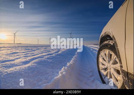 Car tires on winter road covered with snow. Vehicle on snowy country road with wind turbine in the b Stock Photo