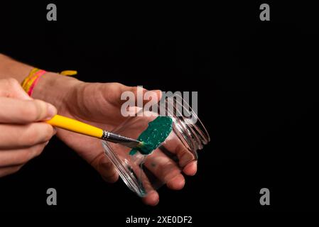 Painters hand holding glass jar and painting green with yellow brush. Isolated on black background. Stock Photo