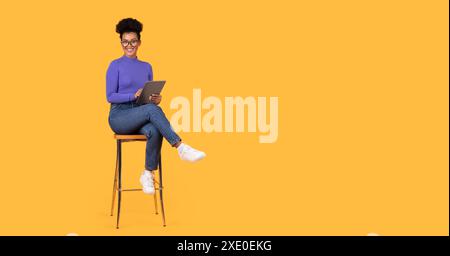 Smiling Woman Using Tablet While Sitting on Stool Against Yellow Stock Photo