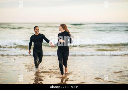 Couple Running Towards The Ocean In Wetsuits After Surf Session Stock Photo
