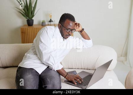 Handsome black African American man working on a laptop computer while sitting on a sofa in a cozy living room. Freelancer working from home Stock Photo