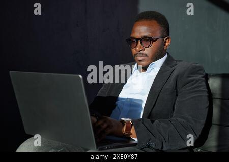 Portrait Of Pensive African Business Man In Glasses Sitting At Desk With Laptop In Home Office, Looking Away With Glad Face Expression. Smiling Guy In Stock Photo