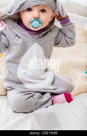 Happy baby girl in cozy pajamas plays with the hood on a soft bed with a smartphone nearby, looking curious and playful with a big smile on her face Stock Photo