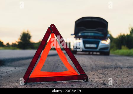 Unrecognizable sad driver in reflective vest. Male driver standing near broken car with open up hood. Red triangle to warn other road users of car bre Stock Photo