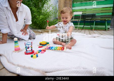Happy baby playing with toys on a blanket outdoors, enjoying playful moments. The child is engaged with colorful educational toys while a smiling woma Stock Photo
