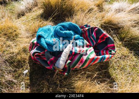 Andean blanket and spinning wheel with alpaca fiber lying in the field surrounded by snowy mountains and blue sky with green and yellow vegetation Stock Photo