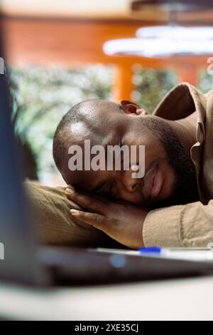 Tired entrepreneur takes a nap on laptop at his home office setup, falling asleep while he works on business management. Man working under pressure and feeling overworked. Stock Photo