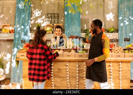 Multicultural people browse locally grown fresh fruits and veggies in greenmarket stands. Black male vendor assisting client at marketplace while female farmer stands behind farm fair booth counter. Stock Photo