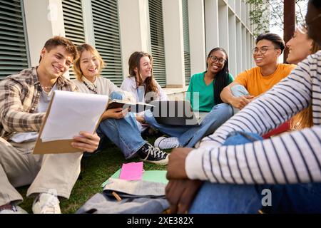Smiling group of young diverse university students people sitting on grass outside faculty building. Stock Photo