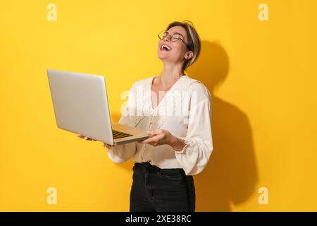 Cheerful woman wearing eyeglasses is using laptop computer on yellow background Stock Photo