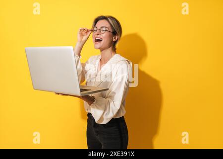 Cheerful woman wearing eyeglasses is using laptop computer on yellow background Stock Photo