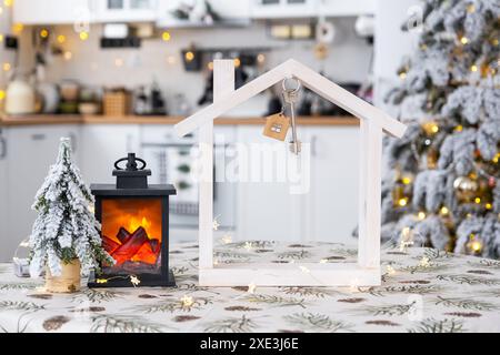 Key and tiny house of a small size on cozy home with Christmas decor on table of festive white kitchen. Gift for New Year. Insur Stock Photo