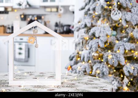 Key and tiny house of a small size on cozy home with Christmas decor on table of festive white kitchen. Gift for New Year. Insur Stock Photo