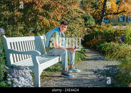 A Teenager Sits On A Bench In The Autumn Park Drinks Coffee From A Thermo Mug And Looks Into A Phone. Portrait Of Handsome Cheer Stock Photo