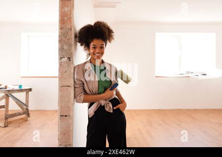 A young attractive African-American woman in casual clothes stands and smiles with a paint roller. Painting the walls. Stock Photo