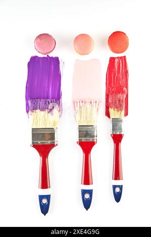 Three paintbrushes with red handles, each dipped in colorful paint, with matching macarons on a white background. Stock Photo