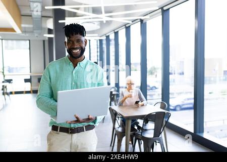 Smiling man holding laptop in modern office space with large windows Stock Photo