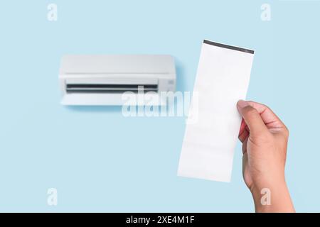 Blank white paper bill holding in a hand with air conditioner blurred on light blue wall in background , Payment concept Stock Photo