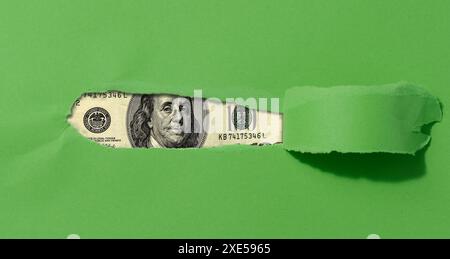 A green sheet of paper with a torn middle and a curled edge, a hundred dollar bill in the hole. Stock Photo