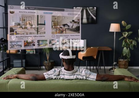 Rear view of unrecognizable Black man wearing VR headset sitting relaxed on sofa in living room surfing Internet Stock Photo