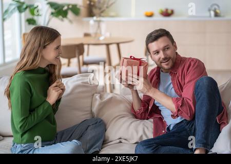 Amazed man, holding gift from loving wife, tries to guess contents of box sitting on couch at home. Stock Photo