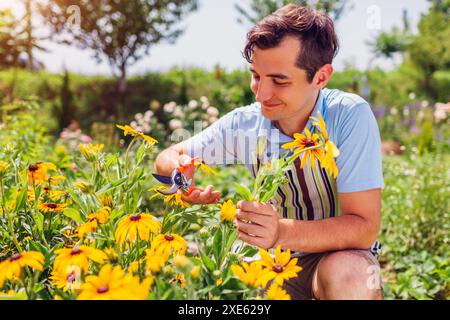 Happy young man picking flowers in summer garden using pruner. Gardener enjoying yellow blooms wearing apron. Leisure outdoor activities Stock Photo