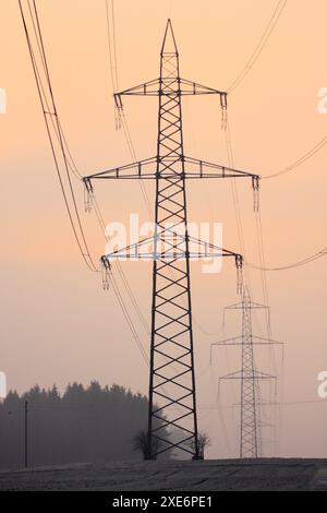 Setting sun behind silhouettes of electricity pylons and power lines, shrouded in orange light amidst fog. Canton of Zurich, Switzerland Stock Photo