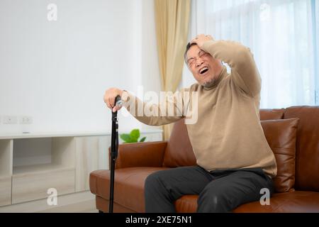 With headache, a senior guy sits on a sofa with a walking stick in his living room at home. Stock Photo