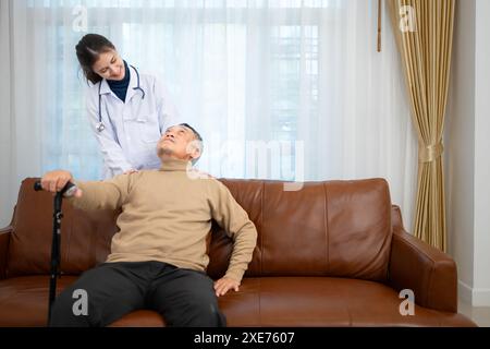 Doctor examines and treats an elderly man seated on a sofa with a cane who is suffering from knee joint pain. Stock Photo