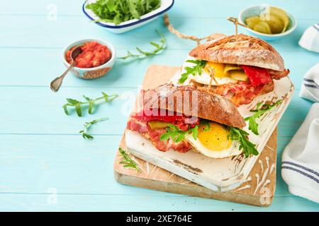 Sandwich layered. English or American freshly prepared picnic sandwich with egg, arugula, bacon, pickles and tomato sause on b turquoise table backgro Stock Photo