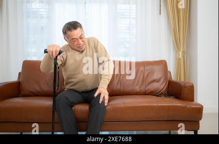 With knee joint pain, a senior guy sits on a sofa with a walking stick in his living room at home. Stock Photo