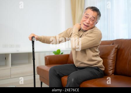 With shoulder pain, a senior guy sits on a sofa with a walking stick in his living room at home. Stock Photo