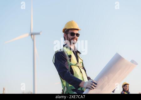 Young man engineer wearing safety helmet and holding blueprint in wind turbine farm. Stock Photo