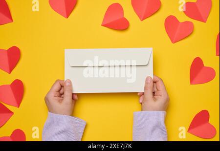 Female hands hold a white rectangular envelope on a yellow background, paper red hearts. Stock Photo