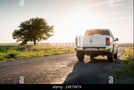 white Volkswagen Amarok on a countryside road in late afternoon. Rear three quarter view of VW pickup truck against the sun, lone tree by the road. Stock Photo