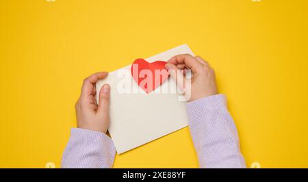 Female hands hold a white rectangular envelope on a yellow background, paper red hearts. Stock Photo
