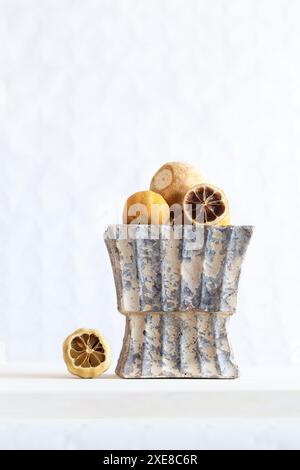 Still life of dried lemons cut into rustic vase on white wooden board on white background. Vertical. Stock Photo